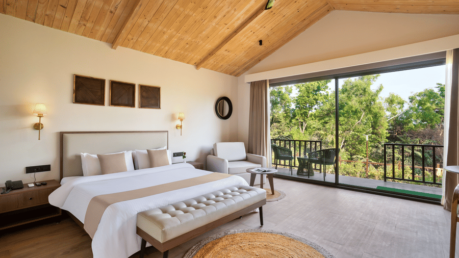An image of a premium cottage featuring a king size bed, a bedside table and lamp on either side, a sofa chair, and a spacious balcony attached to the room at Stone Woods Premier Resort, Gokarna