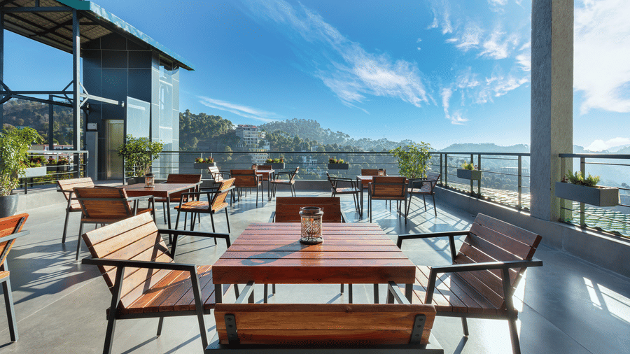Outdoor dining area at Suryavilas Luxury Resort and Spa, featuring wooden tables and chairs on a spacious terrace with panoramic views of the Himalayan hills under a bright blue sky.