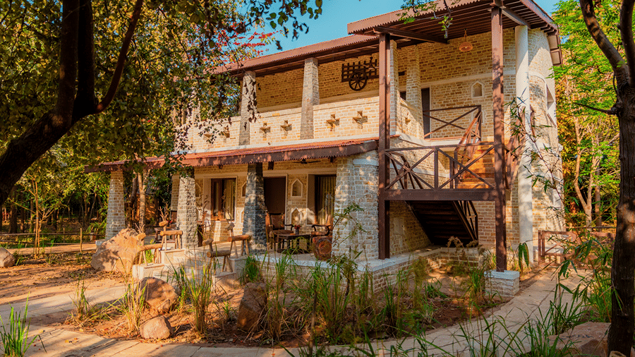 Exterior view of a two-story rustic cottage at Tendu Leaf Jungle Resort & Spa, featuring stone and wood architecture, balcony, ground-floor patio, and surrounding lush foliage.