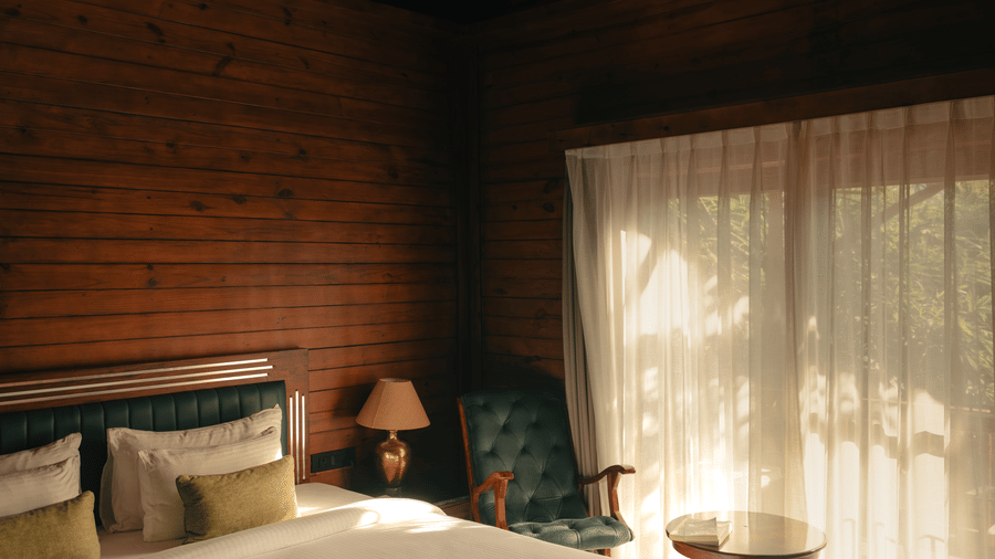 A bedroom at The Ananta Udaipur featuring a king size bed, side table, armchair, small round coffee table, and large window with curtains.