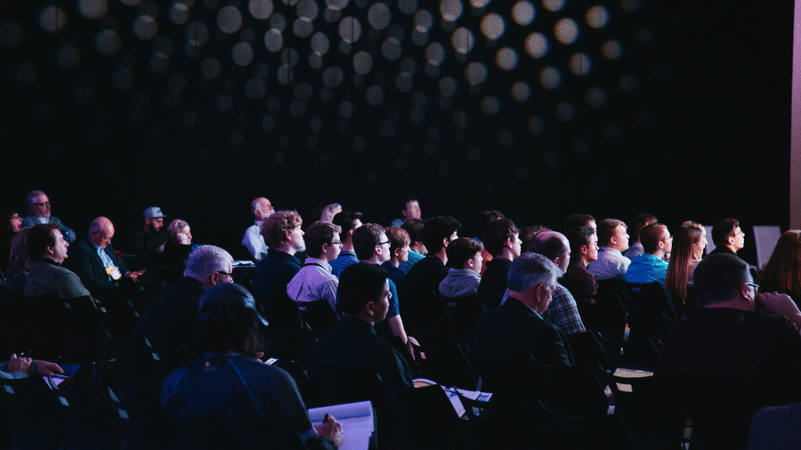 Audience seated in dimly lit room, listening attentively. A backdrop of circular lights adds ambiance. The mood is focused and engaged.