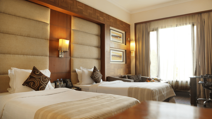 Deluxe Rooms at The Bristol Hotel Gurgaon, Rooms in Gurgaon, featuring twin beds, spreads, pillows, tables, and a window coveed with curtains.