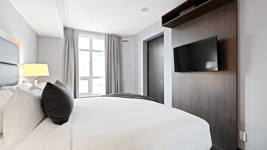 Guest room at The Bristol Hotel, Gurgaon with a king-size bed, grey pillows, a wall-mounted TV on a wood panel, a bedside lamp, and floor-to-ceiling curtains.