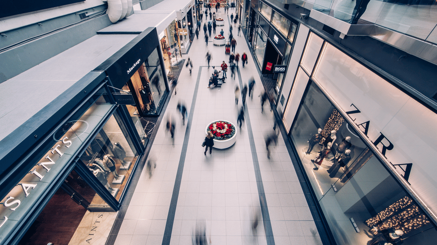 Aerial view of a busy shopping mall corridor with motion-blurred shoppers, festive floral displays, and storefronts including Zara, AllSaints, and Boss.
