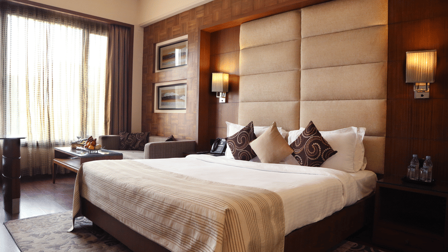 Premium Rooms at The Bristol Hotel Gurgaon, with a large bed, spreads, pillows, lamps, a window, a table and a cushion.