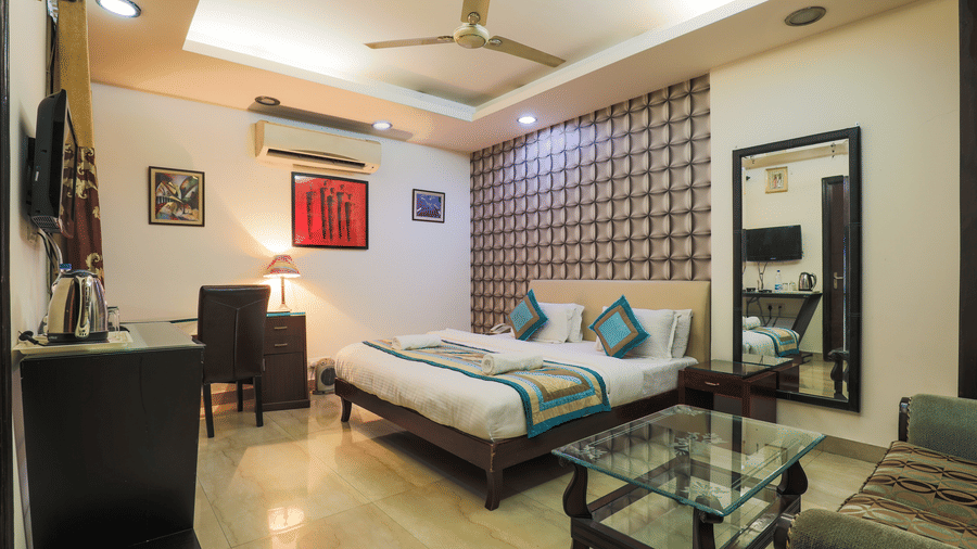 A bedroom featuring a bed, a desk with a chair, a glass table, a sofa, and a mirror that reflects a television.