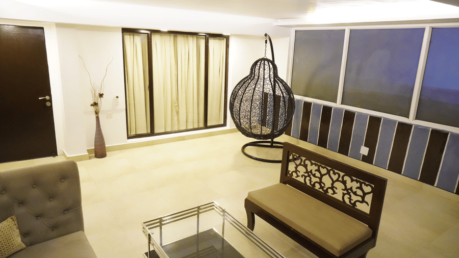 An elevated & wide view of a corridor in Sarovar Portico, Bhubaneswar, featuring an upholstered wooden sofa with a centre table, large glass windows offering views, and a metal hanging chair.