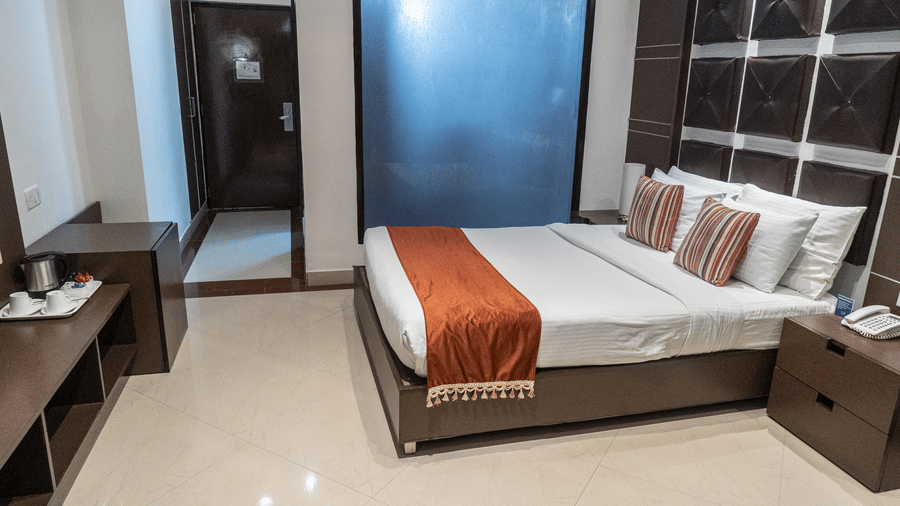 Extended view of the Executive Room of the Sarovar Portico, Bhubaneswar, featuring a plush double bed placed with a cushioned headboard on a wall, beside bedside table, and a large glass frame.