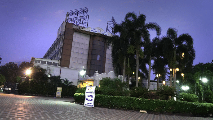 Facade of Sarovar Portico, Bhubaneswar, seen under the dark sky, with a well-lit driveway, a manicured garden, and a board with the name of the hotel on it.