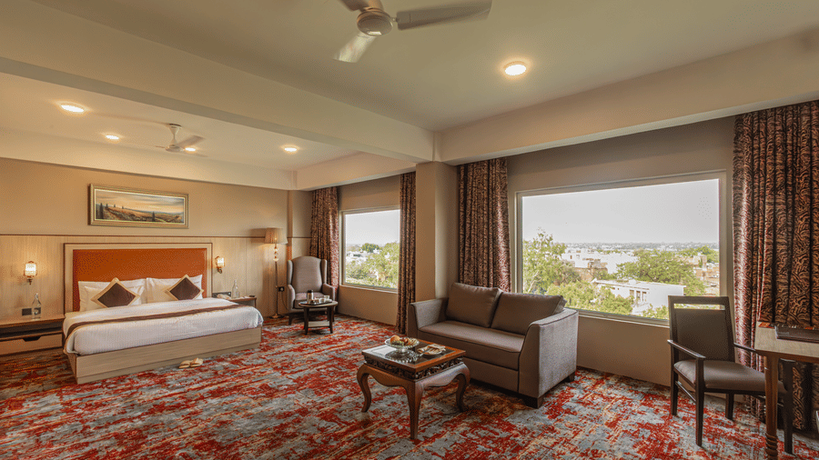 The Junior Suite Room at The Lalita Grand with a double bed, ceiling lights, a 2-seater sofa, ceiling lights, and a window overlooking the greenery.