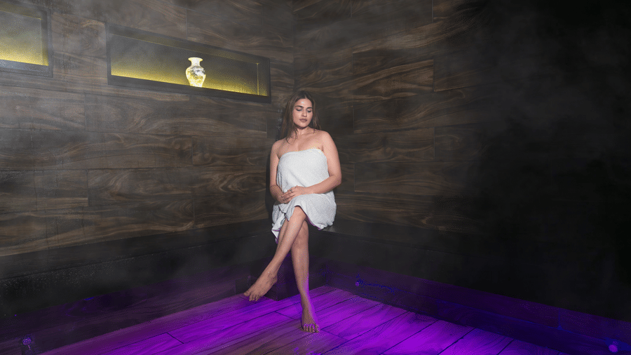 A woman in a bath towel sitting on a counter top of a Sauna Room with smoke filling the room and a vase seen above her on a ledge at  The Manor Luxury Hotel, Shimla.