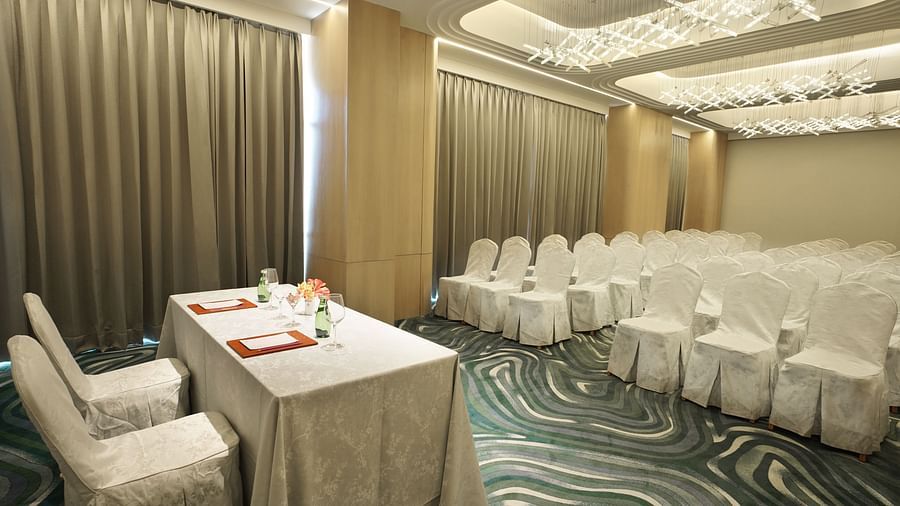 Side view of an event space with rows of white chairs and a white table with orange details. Patterned carpet and draped curtains - The Orchid Hotel, Chandigarh