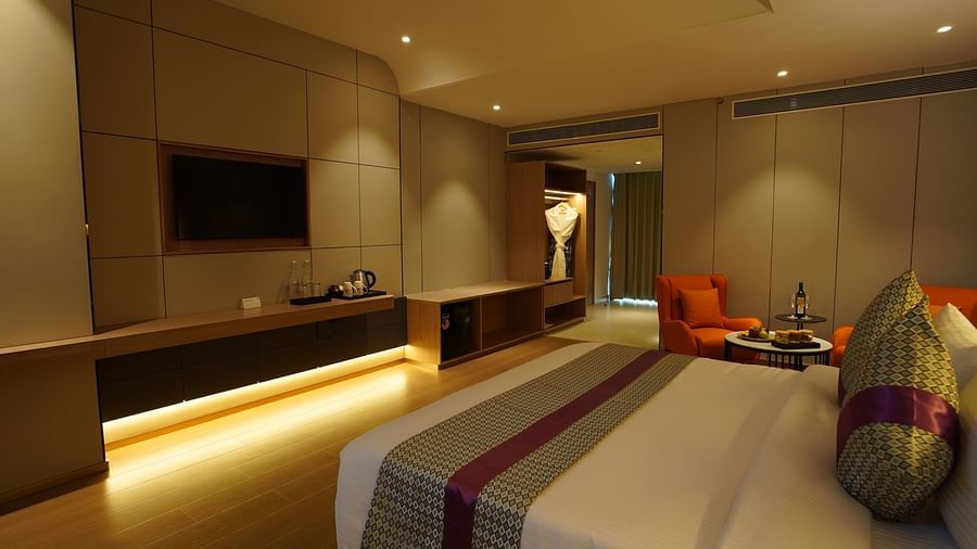 A warmly lit hotel premier room features a king-sized bed with striped accents, a work desk with a TV, and a glimpse into a seating area with orange chairs - The Orchid Hotel, Chandigarh
