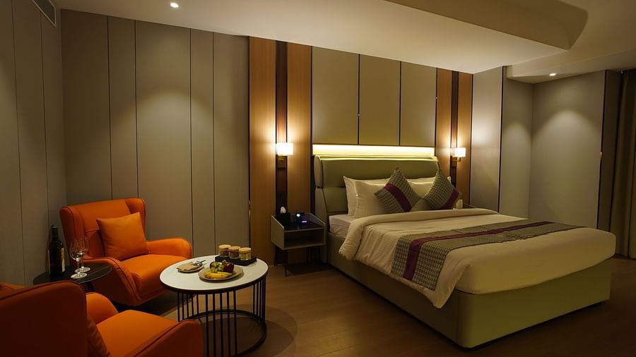 A bright hotel premier room includes a king-sized bed with patterned bedding, a seating area with orange chairs and a round table, and soft lighting - The Orchid Hotel, Chandigarh