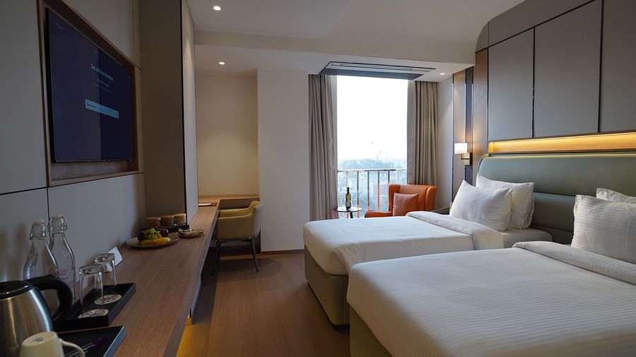 A modern hotel room includes two single beds with white linens, a seating area with a dark sofa, and a view towards a window with city glimpses - The Orchid Hotel, Chandigarh
