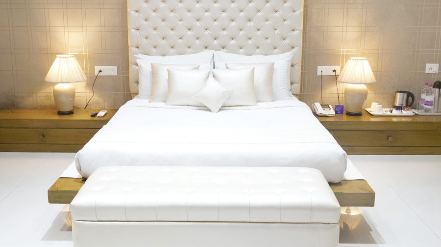 A bedroom in the Suite of Sarovar Portico, Bhubaneswar, seen with ample lighting, a king size bed with upholstered headboard, an upholstered bed bench seen at the foot of the bed, bedside tables, and marble flooring.