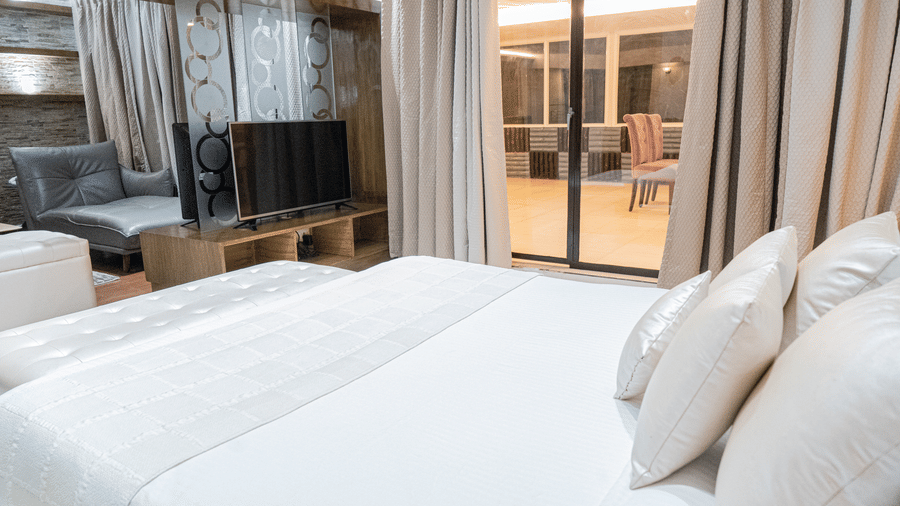 Corner view of the Suite at Sarovar Portico, Bhubaneswar, with the king size bed facing a seating area, a TV placed inside a glass box, and sliding doors leading to the separate seating area situated outside.