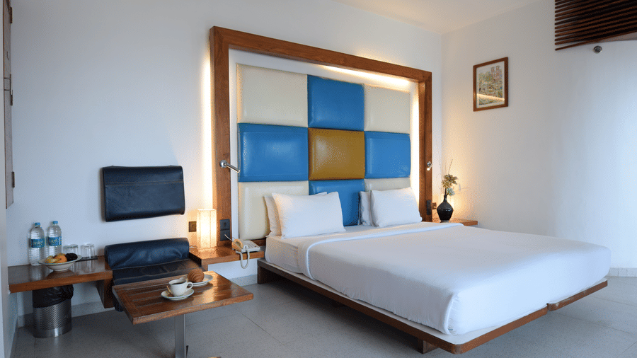 iew of a deluxe room at The Promenade Pondicherry with twin beds, a blue and yellow headboard panel, work desk, wooden flooring, and soft lighting.