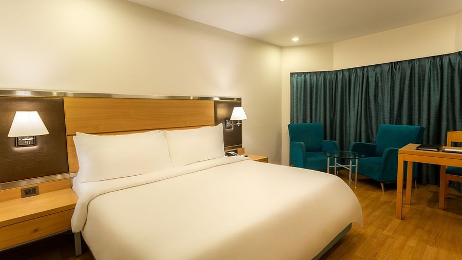 Superior Panoramic Rooms at The Residency Chennai featuring a plush bed with a wooden headboard and lamps on both sides above the nightstands, a sitting space of two couches and a coffee table by the window