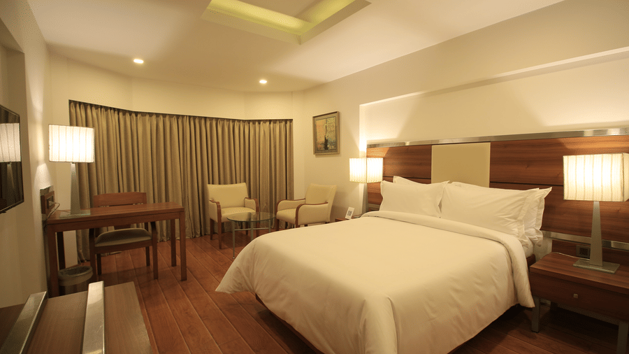 The Superior Rooms at The Residency Chennai, featuring a bed, bedspreads, pillows, chairs, a table, lamps, and windows covered with curtains. 