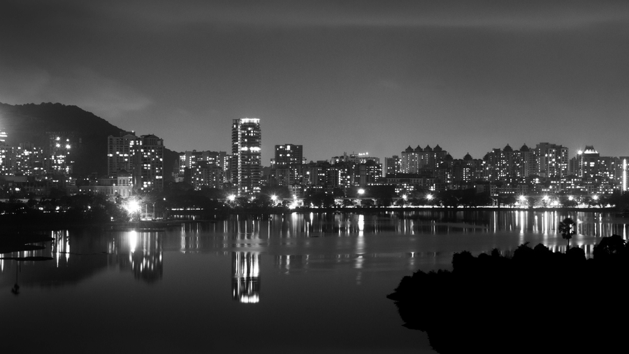 Black and white view of a city skyline at night with illuminated buildings reflected in a calm lake, hills in the background, and dark foreground silhouettes.