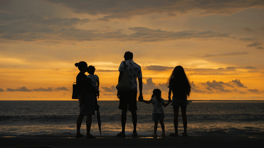 Family of five holding hands on the shore at sunset with dramatic orange and yellow sky reflecting on the water