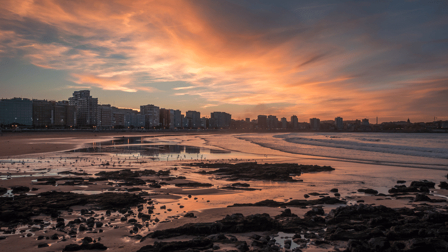 A rocky shoreline with the sun setting and a city skyline in the distance.