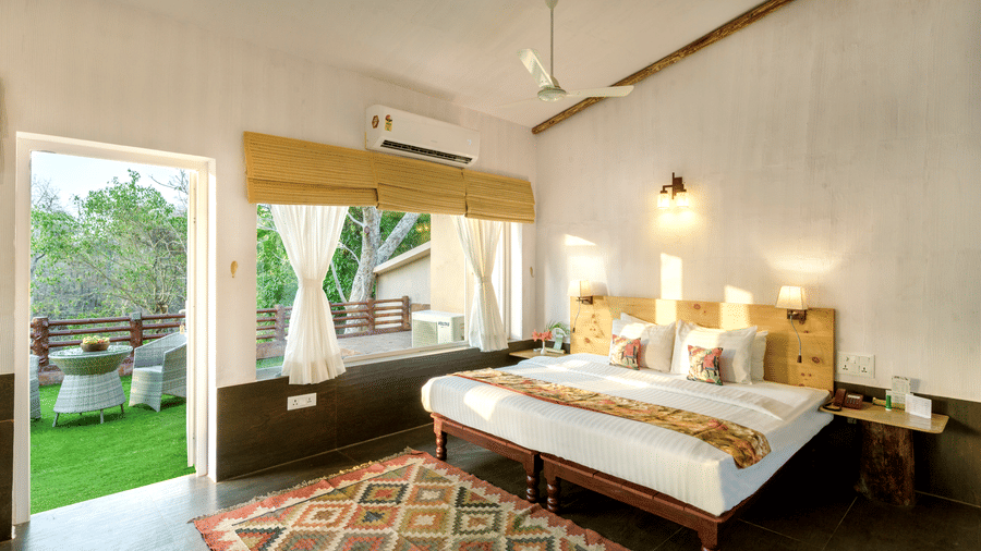 Premier Riverview Suite Twin at The Riverwood Forest Retreat, Pench featuring double size bed, bedside table with telephone, a window with blinds and a curtain, and a balcony with seating area.