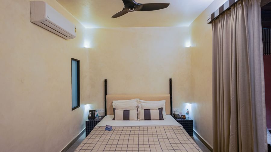 A well-lit photograph showing the elegant simplicity of the Sierra Jacuzzi Suite's bedroom. The comfortable four-poster bed, dressed in crisp white and red bedding, is centered between two bedside tables under a high ceiling with a ceiling fan.