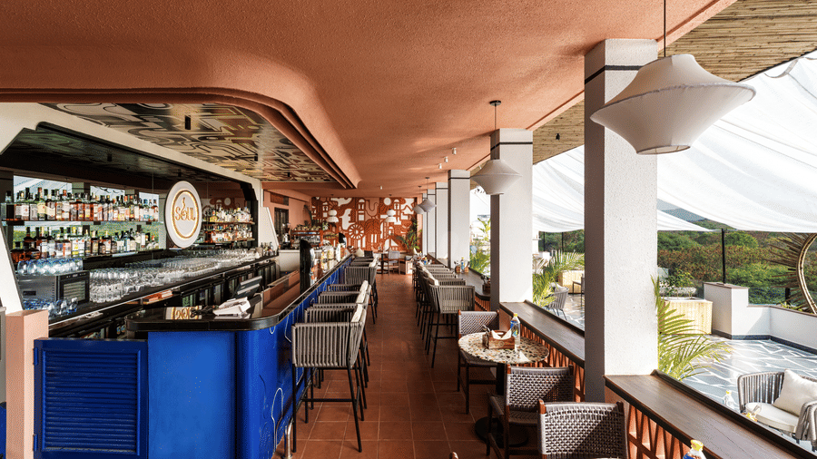 A long rooftop bar counter at The Sierra - By The Lake features stools, small dining tables, and hanging lamps under a shaded structure with open sides.