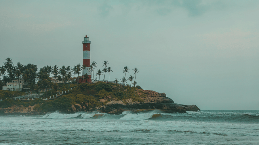 A lighthouse standing on a rocky coastline with waves hitting the shore under a cloudy sky.