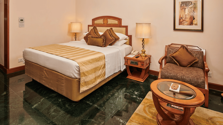 A large bed with pillows, cushions, and bed rugs, beside bed lamps, with a coffee chair, table, and wall decor at The Ummed Ahmedabad.