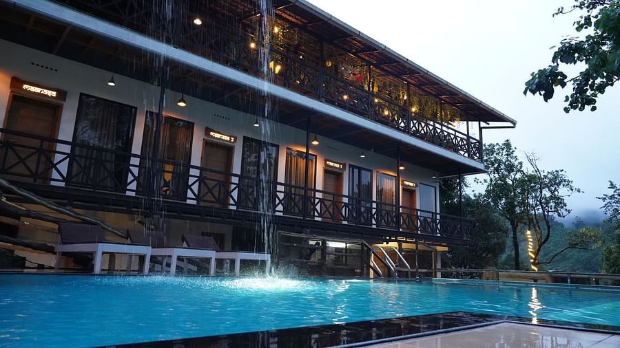 An exterior shot of a modern hotel building with multiple floors and balconies, illuminated by lights, next to a large, inviting swimming pool with blue water. The surrounding appears to be a natural, possibly hilly, landscape. | Premium Pool view suites |