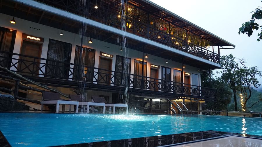 An exterior view of a multi-story hotel building with balconies, illuminated lights, and an infinity pool in the foreground, all set against a natural, possibly misty, landscape. | The Wild Trails Munnar