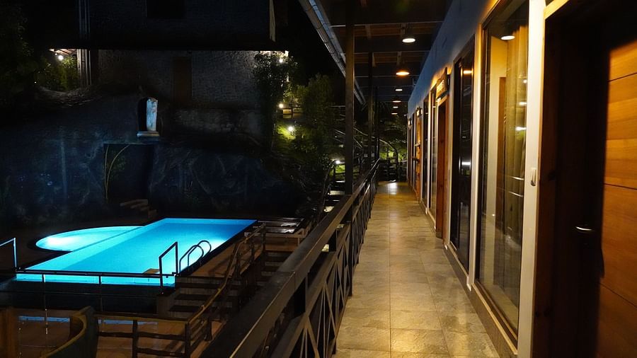 A nighttime view of a long, illuminated walkway alongside a bright blue swimming pool, with the hotel building visible in the background, creating an inviting ambiance. | The Wild Trails Munnar