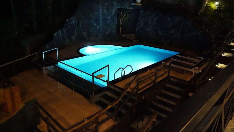 An aerial view of a brightly lit blue swimming pool at night, surrounded by wooden decking and stairs, with dark, rocky and vegetated areas in the background, suggesting a serene and secluded setting. | The Wild Trails Munnar