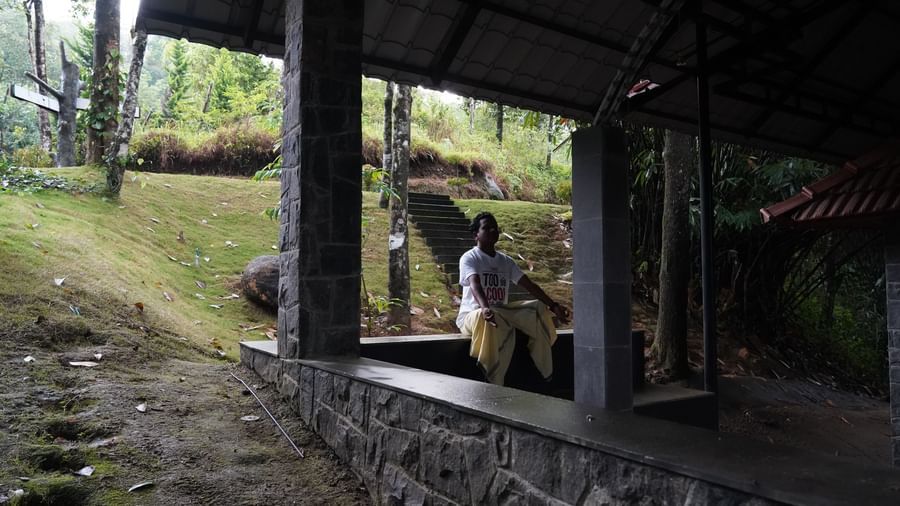 A covered outdoor seating area with a low stone wall and a wooden structure, set within a lush green environment, offering a tranquil space for relaxation. | The Wild Trails Munnar