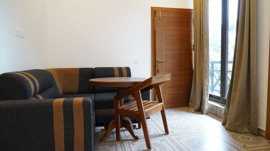 A cozy sitting area in a hotel room, featuring a dark-colored sofa, a small wooden table, and a chair. A doorway leading to a balcony or outdoor area is visible, with natural light coming in. | Premium Pool view suites | The Wild Trails Munnar