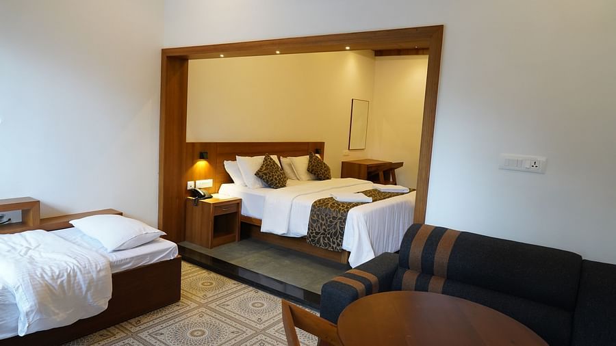 A spacious hotel room featuring a large bed with white linens, a dark-colored sofa, and a round wooden table with chairs. The room has warm lighting and wooden finishes, creating a cozy and luxurious ambiance. | Premium Pool view suites | The Wild Trails M