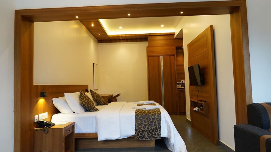 A luxurious hotel room featuring a comfortable bed with white linens, wooden wall panels, and elegant lighting fixtures. There's also a glimpse of a sitting area with a lamp. | Premium Pool view suites | The Wild Trails Munnar