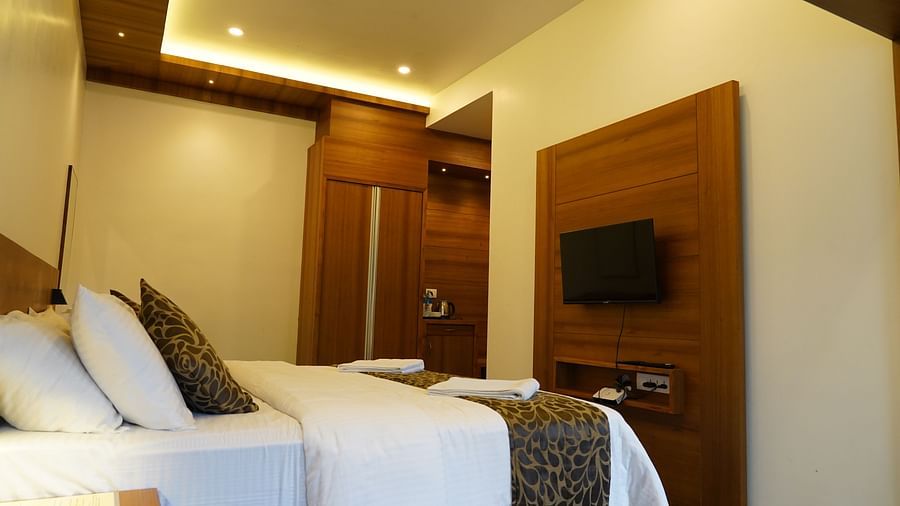 A view inside a well-appointed hotel room, showcasing a comfortable bed with crisp white and patterned linens, a wooden wall panel behind the bed, and a mounted television. The lighting is warm and inviting. | Premium Pool view suites | The Wild Trails Mun