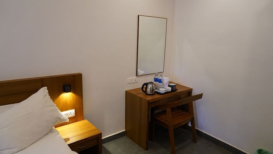 A simple yet comfortable hotel room featuring a bed with a wooden headboard, a bedside table with a lamp, and a wooden desk and chair. A mirror hangs on the wall, and bottled water is provided on the desk. | Premium Pool view suites | The Wild Trails Munna