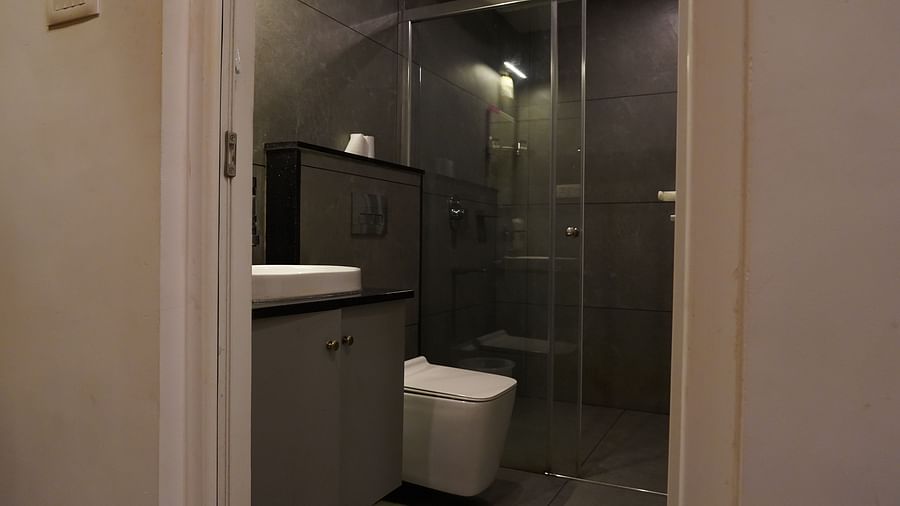 A hotel bathroom with a sleek design, featuring grey tiled walls, a white basin, a wall-mounted faucet, and a large mirror. There's also a shower area visible through a glass partition. | Premium Pool view suites | The Wild Trails Munnar