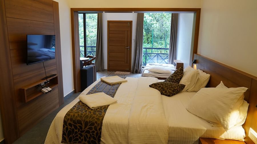 A hotel room with warm lighting, featuring a bed with white and patterned linens, a wooden headboard, and a mounted television. A large glass door or window leads to an outdoor view | Premium Pool view suites | The Wild Trails Munnar