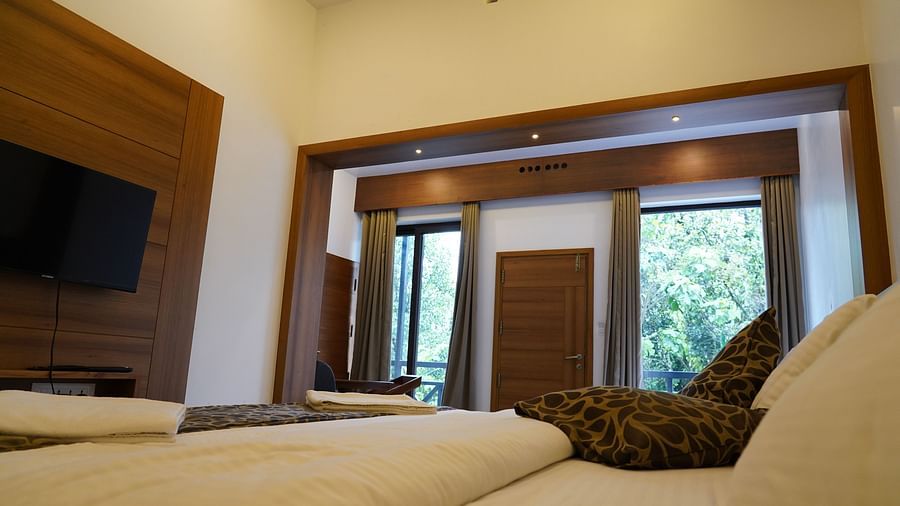A hotel room with warm lighting, featuring a bed with white and patterned linens, a wooden headboard, and a mounted television. A large glass door or window leads to an outdoor view | Premium Pool view suites | The Wild Trails Munnar