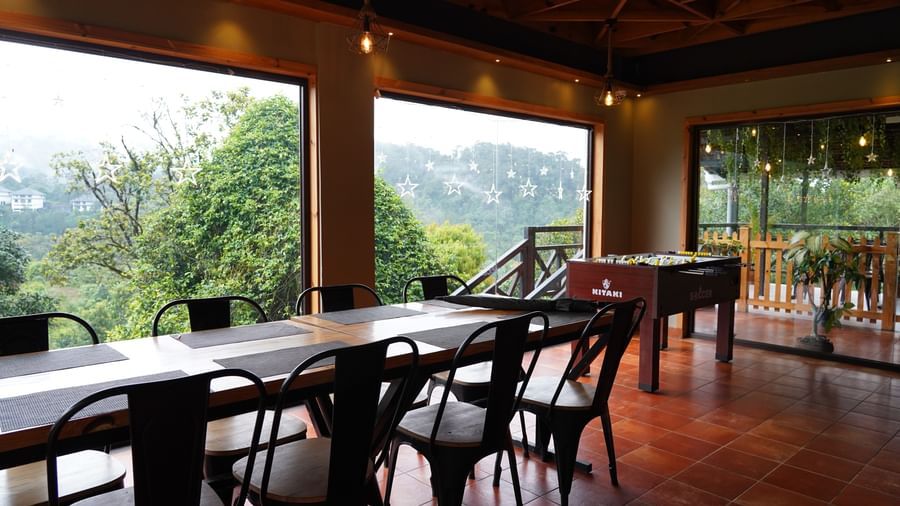 A spacious indoor dining or common area with long wooden tables and black chairs, featuring large windows that offer expansive views of the misty, green mountainous landscape outside. The floor is tiled, and the ceiling has wooden beams. | The Wild Trails 