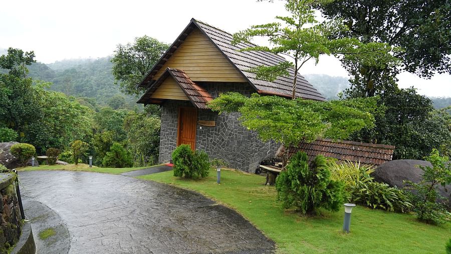 A traditional-style building with a distinctive roof is set amidst lush green hills with a wet road. | Premium Forest View Cottage With Balcony | The Wild Trails Munnar