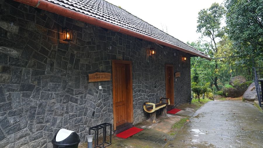 An exterior view of a stone-clad building with a tiled roof, set amidst green trees. | Premium Forest View Suite with Balcony | The Wild Trails Munnar