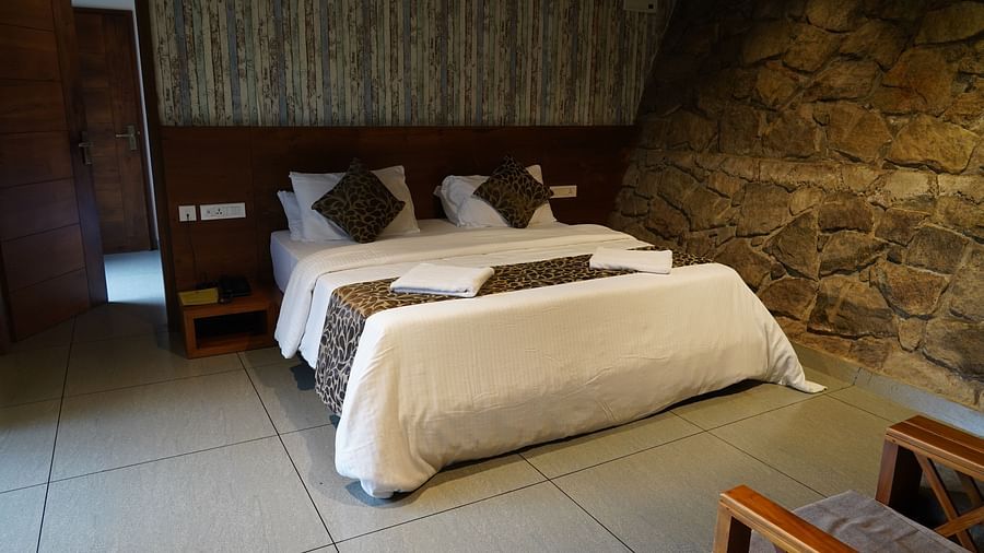 A bedroom with a bed, white linens, and a stone-textured accent wall, showcasing wooden furniture. | Premium Forest view 2 Bedroom Interconnecting Suite Cottage with Balcony | The Wild Trails Munnar