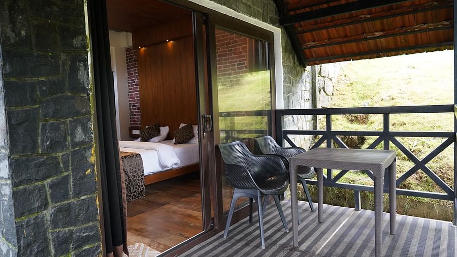 A room with a bed seen through a glass door, leading to a balcony with a table and chairs. | Premium Forest View Cottage With Balcony | The Wild Trails Munnar